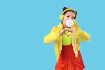 Trendy girl blowing bubble gum while making heart with hear hands on blue background