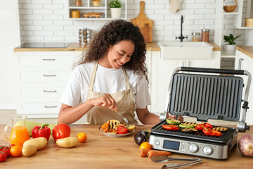 Beautiful young happy African-American woman eating tasty vegetables cooked on modern electric grill in kitchen