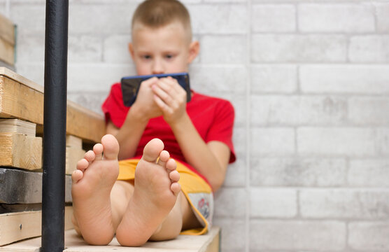 The boy's feet are close, playing on the phone. A child sitting on the steps of a new house. Solitude. Addiction to the internet.