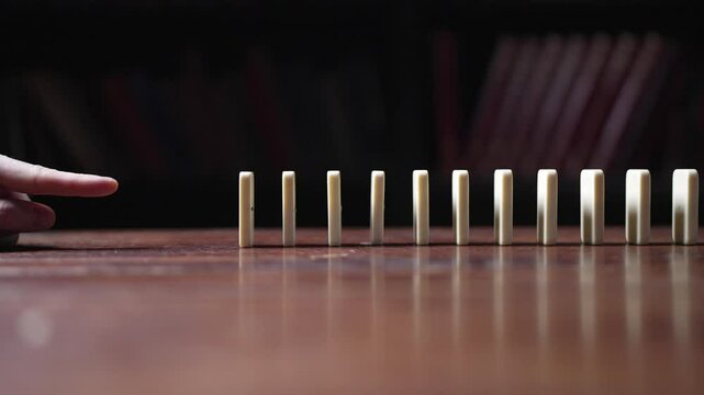 A finger pushes a line of white dominos, triggering a chain reaction in slow motion and showcasing the domino effect