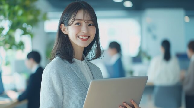 Smiling Japanese businesswoman standing in an office, holding her laptop. She is wearing casual clothes, and other people working behind her are blurred.,generated ai.  
