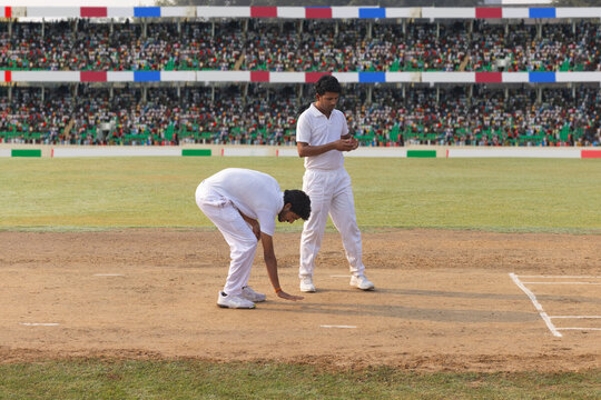 A bowler marks their run-up while a captain stands behind them during a cricket game