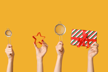 Female hands holding sieves with Christmas gift and cookie cutter on yellow background