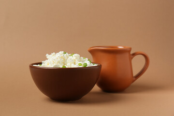 Bowl of fresh cottage cheese with green onion and jug of milk on brown background