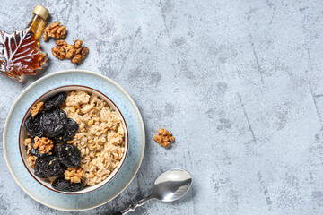 Bowl of tasty oatmeal with prunes, walnuts and bottle of maple syrup on grey grunge background