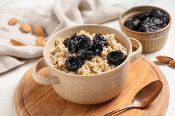 Wooden board with pot of tasty oatmeal and prunes on light background