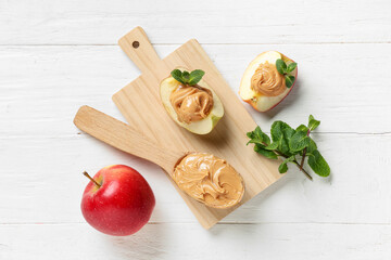 Board with tasty apple wedges, nut butter and mint leaves on white wooden background