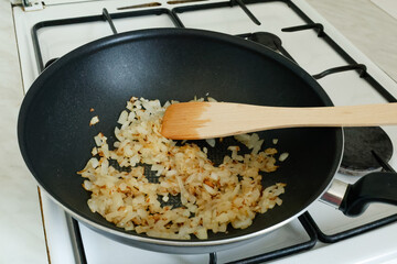 A frying pan with chopped onions being stirred with a wooden spatula on a gas stove. 