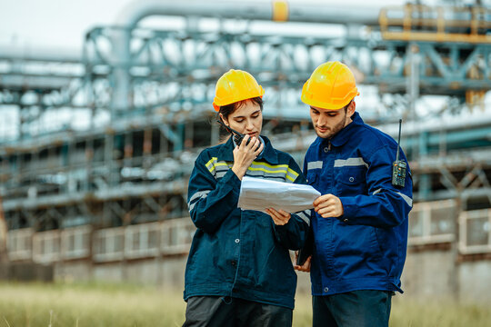 Workers review plans at industrial site while wearing safety helmets and communicating with radios
