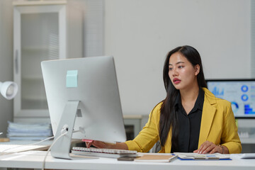 Professional woman in a yellow blazer working at a desk, analyzing data on a computer screen in a...