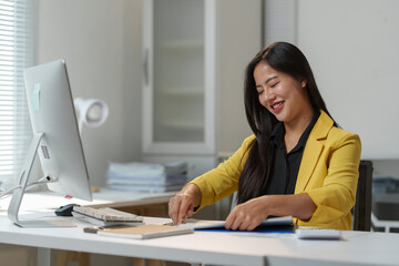 Professional woman in a bright office setting, organizing paperwork at her desk. She is focused and smiling, suggesting a positive work environment and efficient productivity