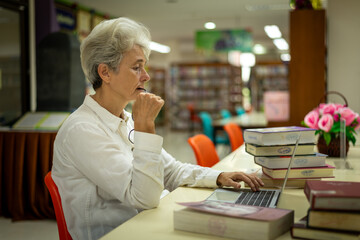 Elderly woman sitting in a library, combining traditional and modern approaches to learning or research by book and laptop computer sense of purpose dedication to personal grown concept.	