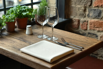 Rustic wooden table with glasses, cutlery, and greenery, creating a cozy dining setting near a window.