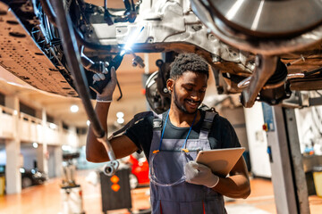Smiling mechanic inspecting car's undercarriage with digital tablet in auto repair shop