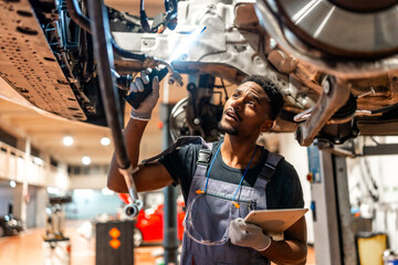 Mechanic inspecting car chassis with flashlight and tablet in workshop