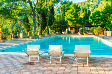 three white desk chairs or chaise lounge with blue swimming pool and hotel green garden on backgeound of vacation summer landscape
