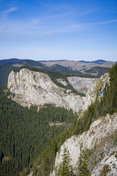 A Majestic Mountain Landscape Showcases Rocky Cliffs, Dense Green Pine Forests, Rolling Hills, And A Clear Blue Sky In The Background