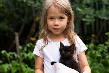A cute little child girl looking at camera holding a bowl with a black kitten outdoors in nature