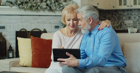 Happy senior old couple laughing talking relaxing on sofa using digital tablet, cheerful mature aged family spending time with gadget having fun. - Powered by Adobe