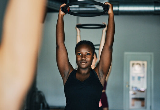 Fit young black woman in sportswear working out with a ring while kneeling on a reformer during an exercise class with other women at a pilates gym