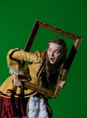 Fototapeta premium Woman in ancient outfit on chroma key green screen background. Female in renaissance dress holding antique frame and posing as portrait painting at camera.