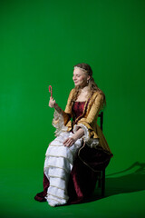 Woman in ancient outfit on chroma key green screen background. Female in renaissance dress sitting holding and looking at candy cane in hand.