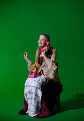 Woman in ancient outfit on chroma key green screen background. Female in renaissance dress sitting and talking on pink retro wired phone, smiling expression.
