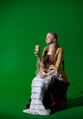 Woman in ancient outfit on chroma key green screen background. Female in renaissance dress sitting and holding coffee in paper cup, smiling.