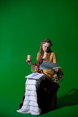 Woman in ancient outfit on chroma key green screen background. Female in renaissance dress sitting reading paper documents with infographic and holding coffee cup.