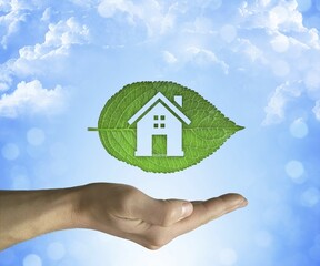 Opened hand holding a green leaf with a house icon inside on a blue sky background. Eco house concept