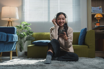 Obraz premium Smiling young woman sitting on a cozy carpet, listening to music with headphones and using a smartphone in a modern living room. Relaxed atmosphere with stylish decor and natural light