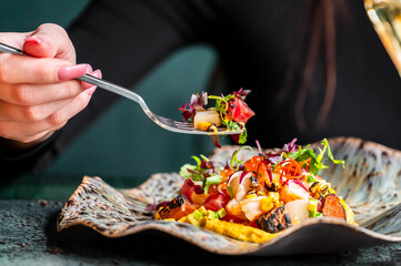 A close-up of a hand holding a fork with a colorful salad featuring fresh vegetables, herbs, and grilled ingredients on a stylish plate. Vivid colors create an appetizing presentation.