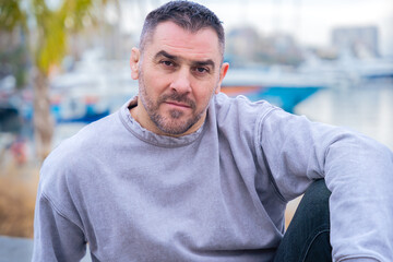 Fototapeta premium Relaxed man wearing a gray sweatshirt sitting outdoors near a marina, looking confidently at the camera. The calm setting and soft lighting enhance his thoughtful expression. 