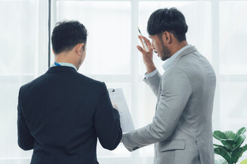 Business Discussion by Window: Two businessmen in suits engage in a serious discussion, viewed from behind, as they review documents near a bright window.