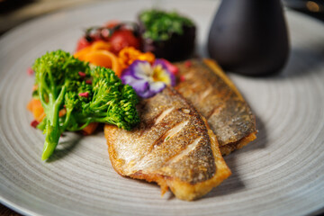 Crispy fish fillet paired with fresh broccoli, cherry tomatoes, and carrot ribbons, topped with colorful edible flowers. Served with a dark sauce jug. Elegant and healthy meal presentation