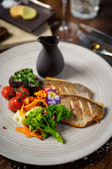 Pan-seared fish fillet with vibrant vegetables, including broccoli, cherry tomatoes, and carrot ribbons, garnished with edible flowers. Accompanied by a dark sauce jug. Fine dining closeup