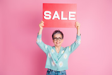 Mature woman holding a sale sign on a pink background, expressing joy and showcasing advertising and shopping concepts