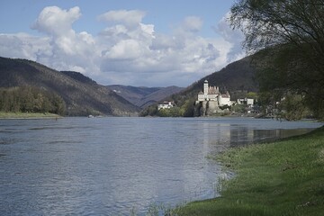 View of a calm river with a castle on the bank, surrounded by wooded mountains and a cloudy sky Schönbühel on the Danube World Heritage Site Wachau Lower Austria
