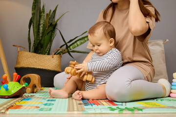 A joyful baby plays with wooden toys on a colorful play mat, seated next to a parent in a stylish and serene room with plants. The child explores and interacts with the toys