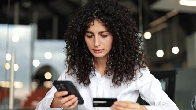 Confused businesswoman with curly hair makes online payment on smartphone using credit card in modern office. She has payment problem.
