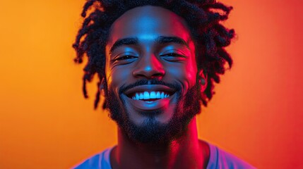 A young man with dreadlocks smiles brightly at the camera, bathed in colorful neon lighting.