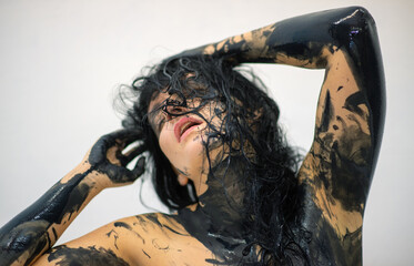 Sexy woman covered in black liquid paint poses expressively, her wet hair partially veiling her face. The intense gaze and raw texture create a bold, artistic, and sensual atmosphere