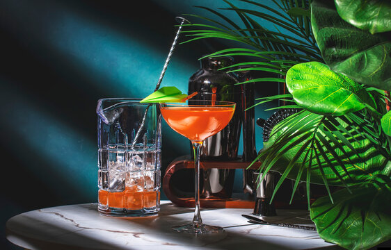 Paper plane alcoholic cocktail drink with bourbon, orange bitters, amaro and lemon juice. Marble table, blue background with tropical leaves