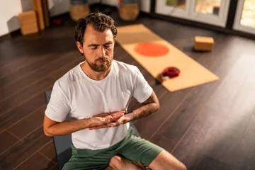 Fototapete Bearded man meditating in lotus position on yoga mat, practicing mindfulness and enjoying peace at home, promoting mental health and relaxation through yoga exercises © DusanJelicic