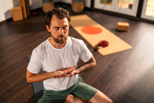 Bearded man meditating in lotus position on yoga mat, practicing mindfulness and enjoying peace at home, promoting mental health and relaxation through yoga exercises