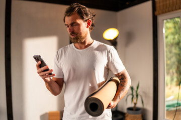 Bearded man holding a yoga mat while using a smartphone, preparing for an online class or...