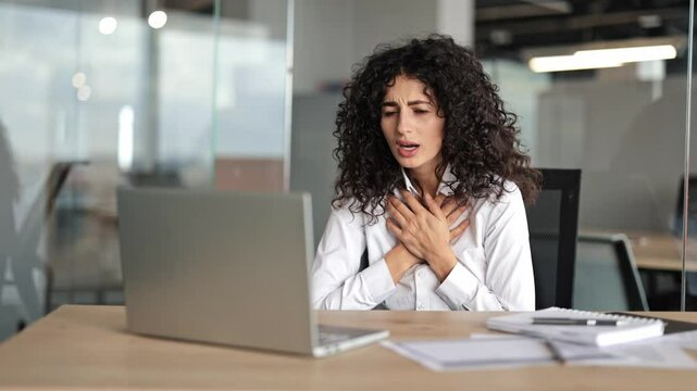 Curly haired businesswoman in modern office has heart attack or anxiety. Shocked female has breathing problems at work. Woman works with laptop.