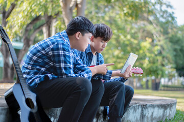 Fototapeta premium Asian teenaged boy spending his free times with his friend on weekend by practising and playing acoustic guitar or ukulele in the park together, recreational activity of teenagers concept.