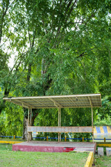 An old outdoor shelter with wooden benches surrounded by lush green trees.
