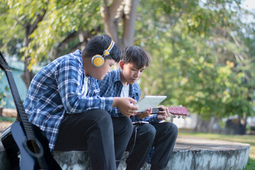 Fototapeta premium Asian teenaged boy spending his free times with his friend on weekend by practising and playing acoustic guitar or ukulele in the park together, recreational activity of teenagers concept.
